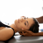 A woman lays on a padded table while a chiropractor adjusts her head to the side