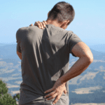 A man standing at a scenic lookout places a hand on his lower back in pain.