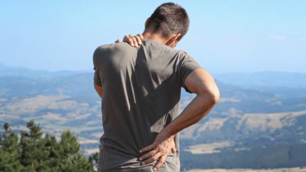 A man standing at a scenic lookout places a hand on his lower back in pain.