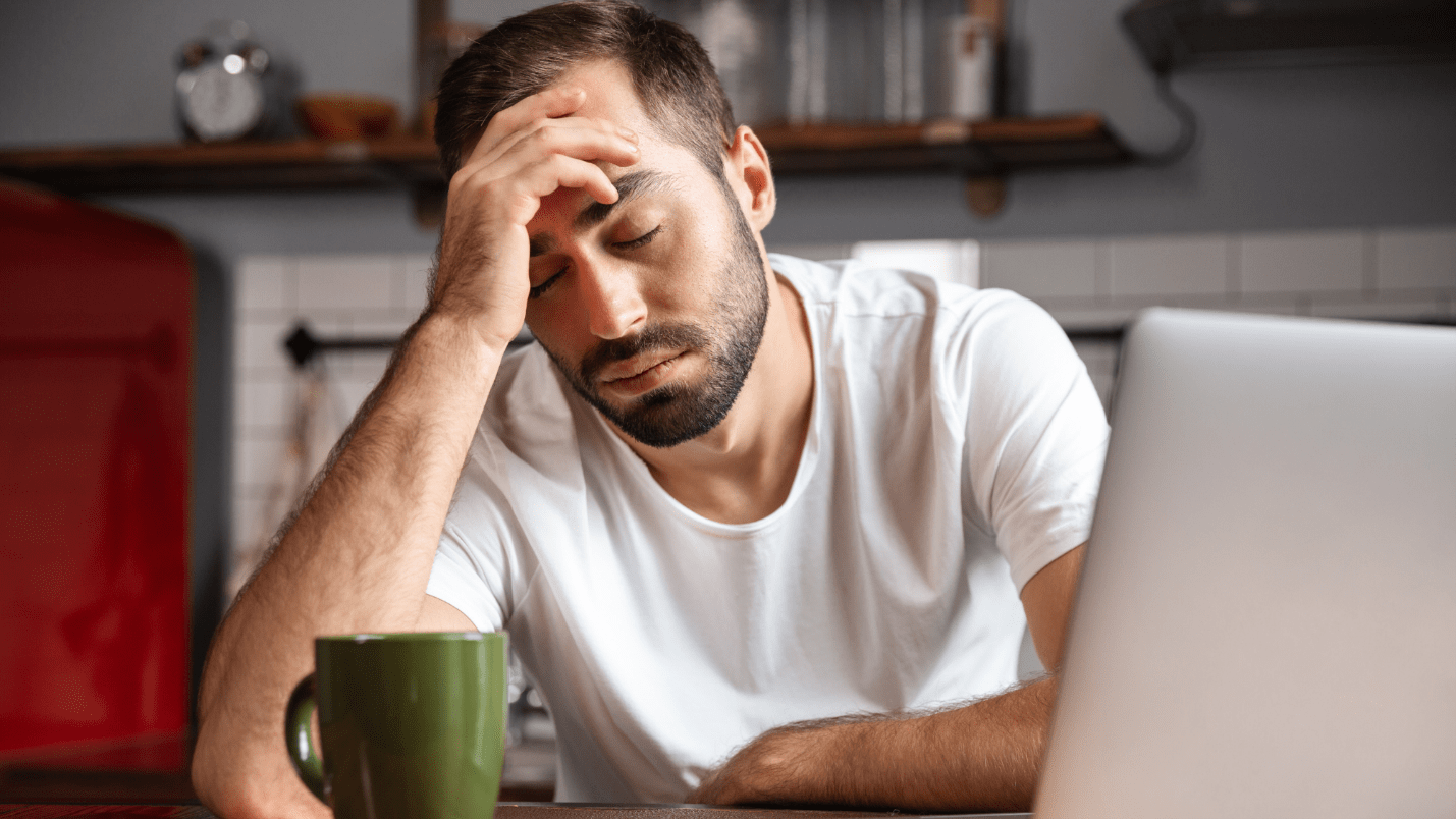 A tired man rests his head on his hand, in front of him is a laptop and a cup of coffee.