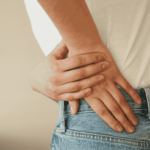 A person in jeans and a t-shirt touches their lower back, in pain.