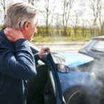 A man exits a car after a collision, hand on the back of his neck.