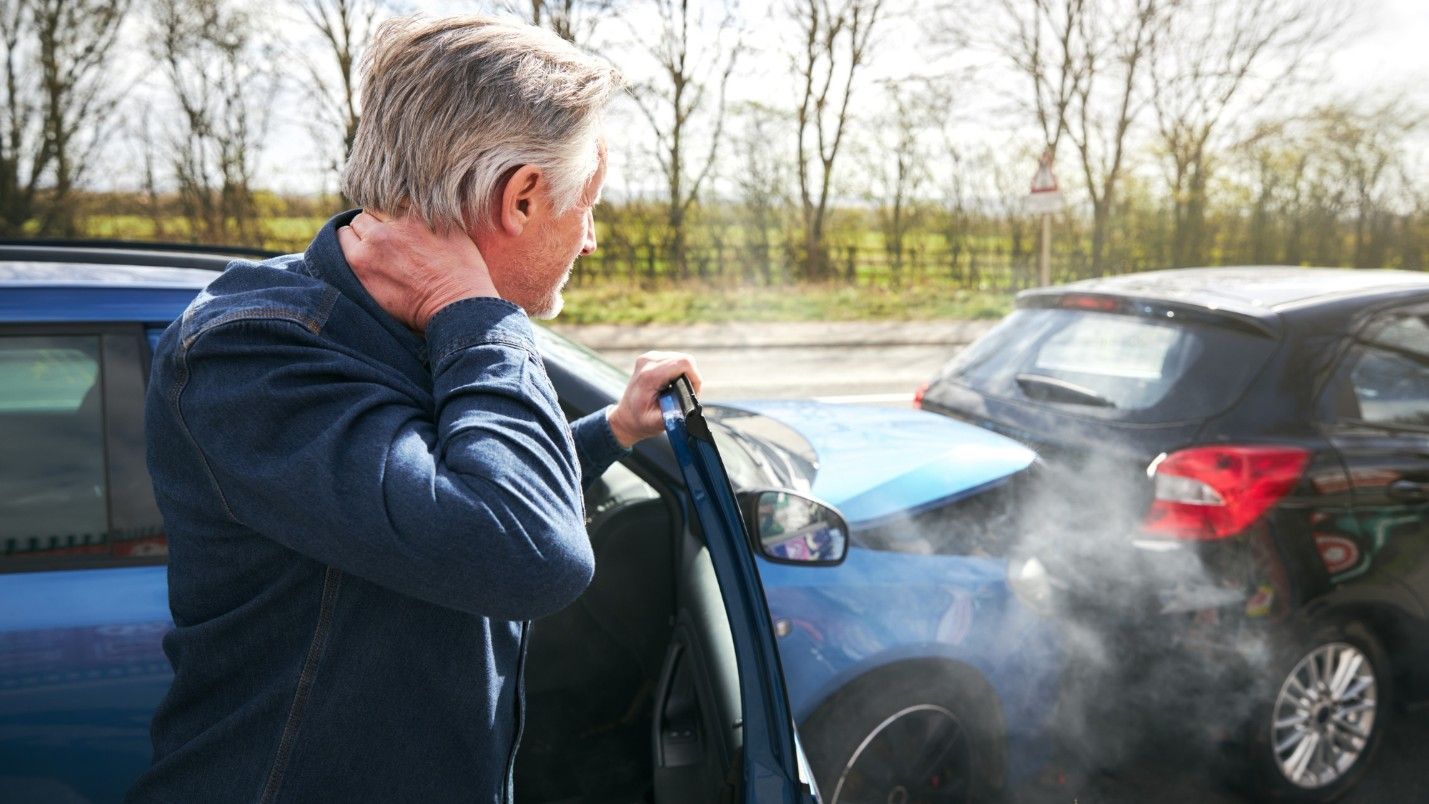 A man exits a car after a collision, hand on the back of his neck.