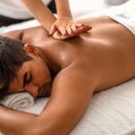 A man lays on a massage table, a masseuse kneads into his back.