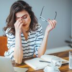 A woman in front of a laptop pinching the bridge of her nose, stressed out.