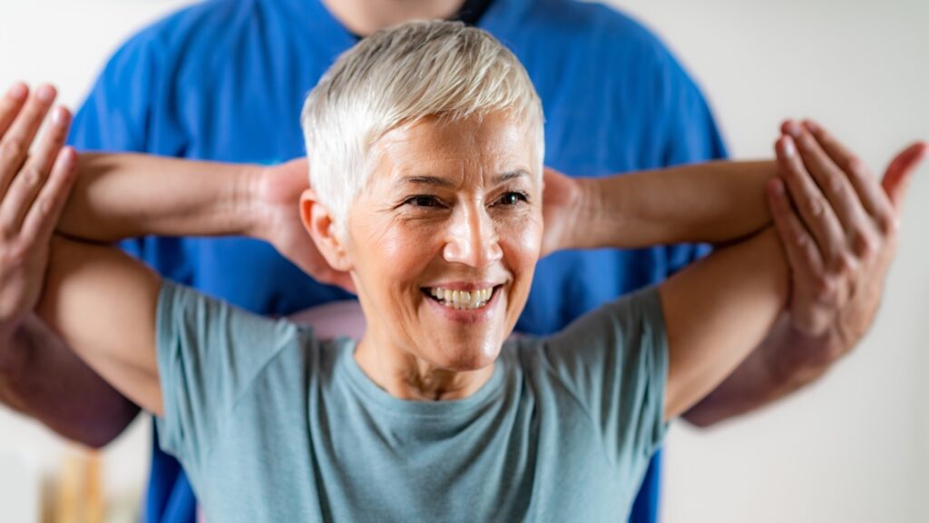 An older woman holds her arms behind her head during chiropractic adjustment.